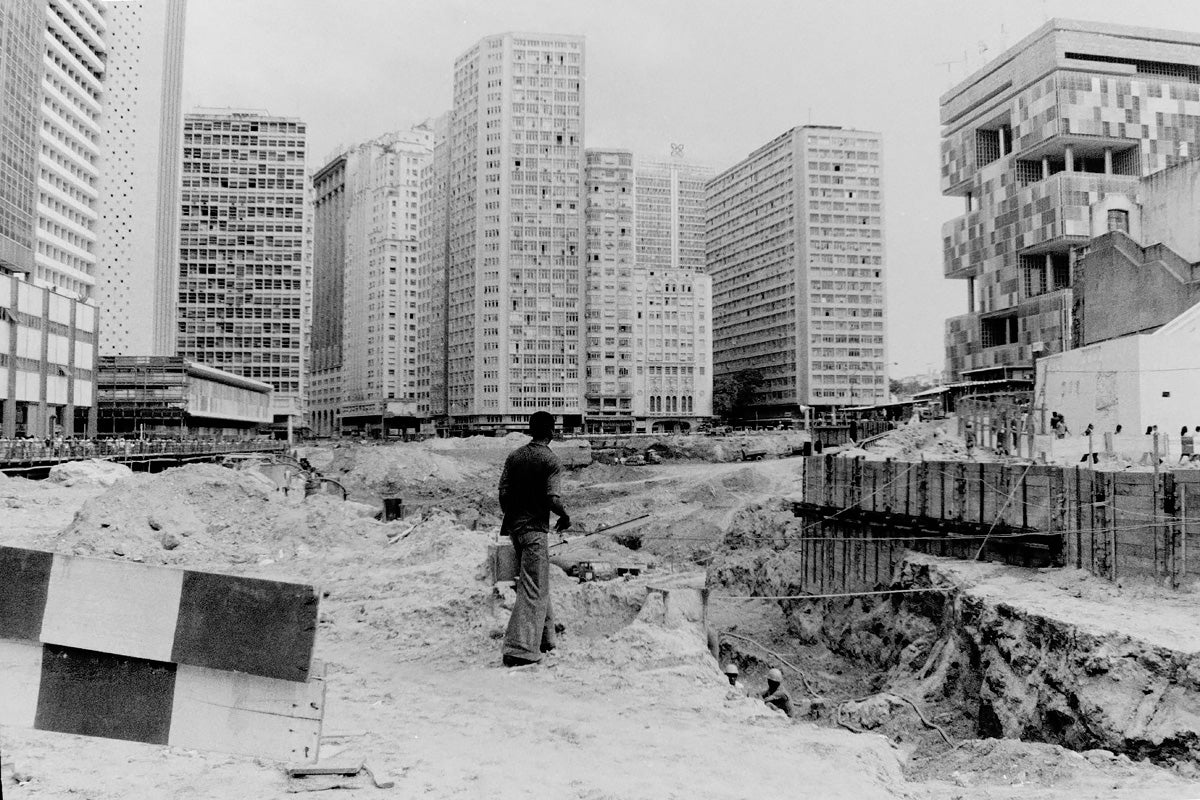 Black and white photograph of Rio de Janeiro. View of man's back. In the background, many tall buildings; in the front, the construction of the Carioca subway station.